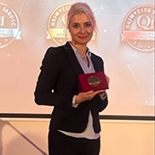 Uśmiechnięta kobieta w garniturze trzyma w rękach medal, stojąc na tle transparentów z napisami "Najwyższa Jakość QI".