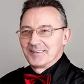 Mężczyzna w okularach i czerwonej muszce uśmiecha się, patrząc lekko w lewo. Tło jest jasne z subtelnym, geometrycznym wzorem.